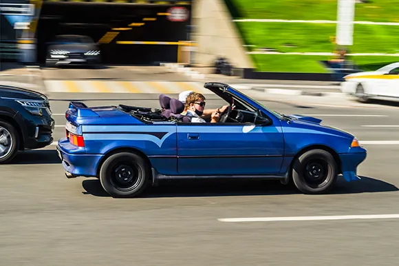 A blue Geo Metro Convertible and its passengers speeding on the highway.